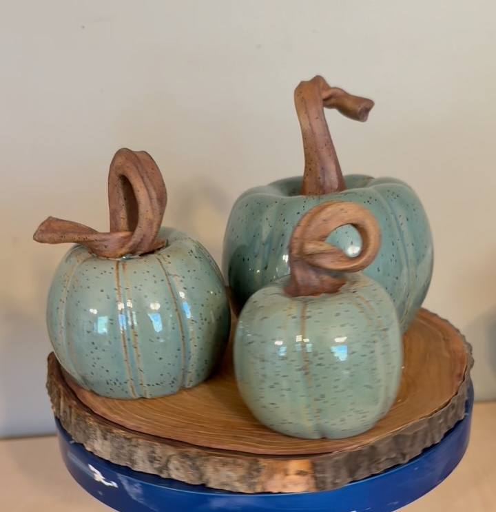 Three celadon-green stoneware pumpkins with twisting clay-brown stems on a wood slice