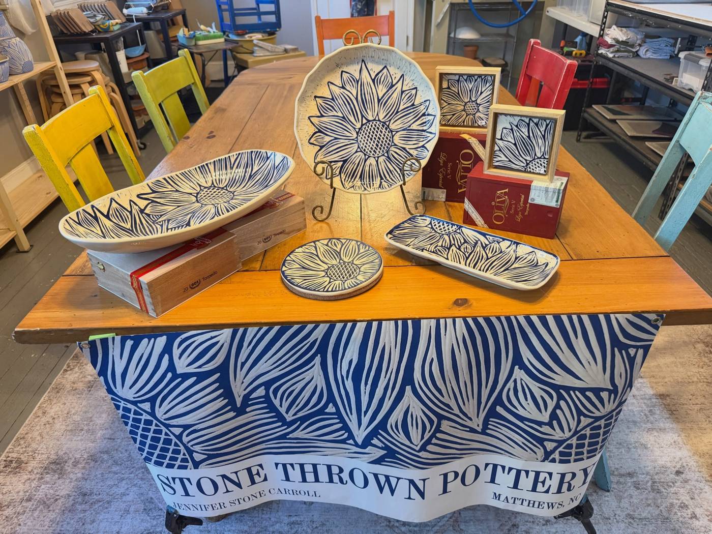 A market table of carved sunflower platters and trays over a Stone Thrown Pottery banner